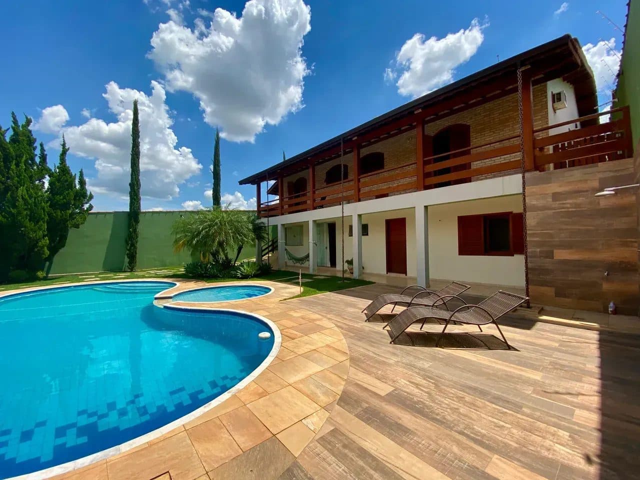 Piscina com formato orgânico, deck de madeira e espreguiçadeiras, vista para a fachada da casa.