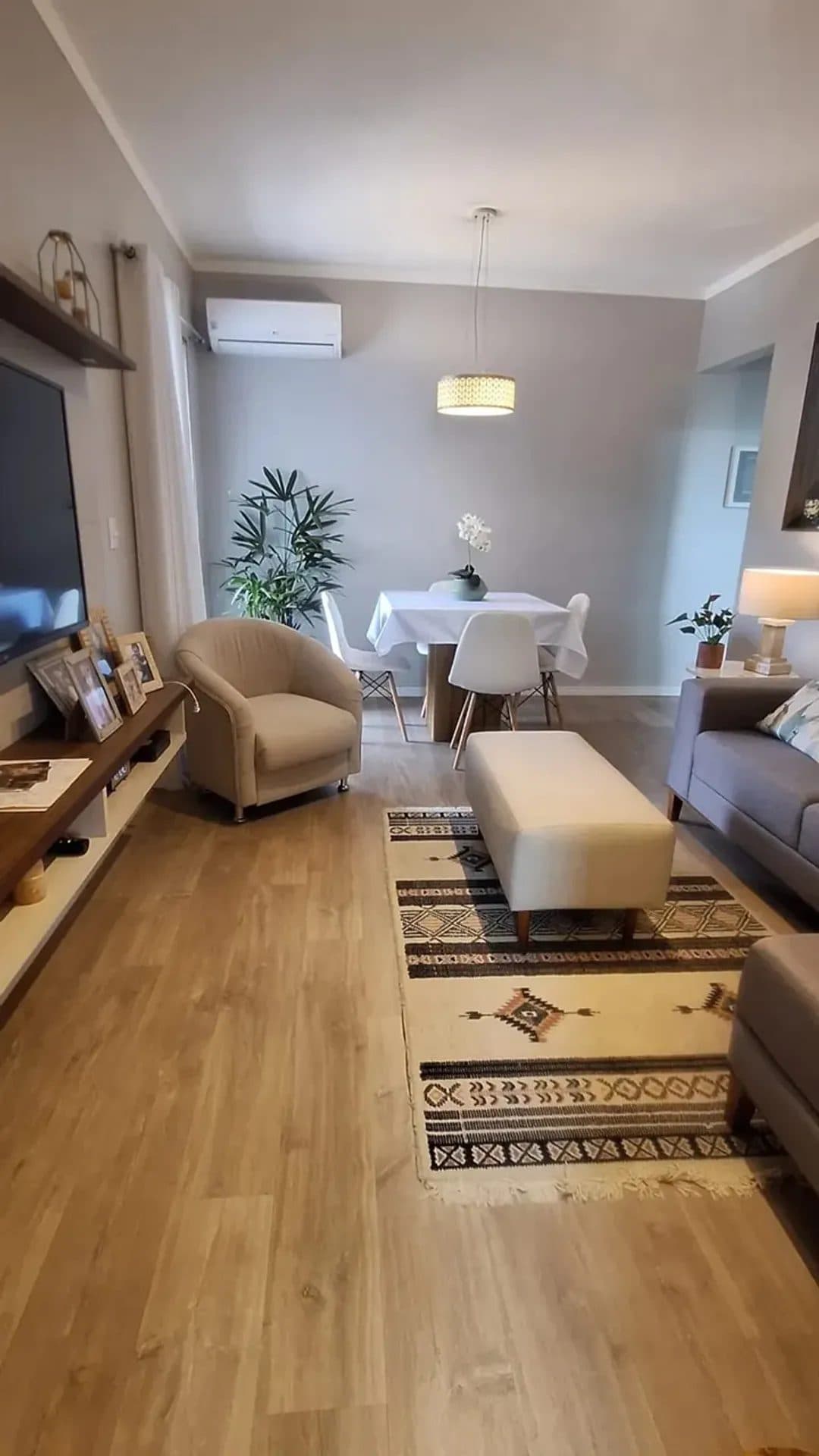 Sala de estar com painel de TV, sofá cinza, tapete geométrico e mesa de jantar ao fundo.