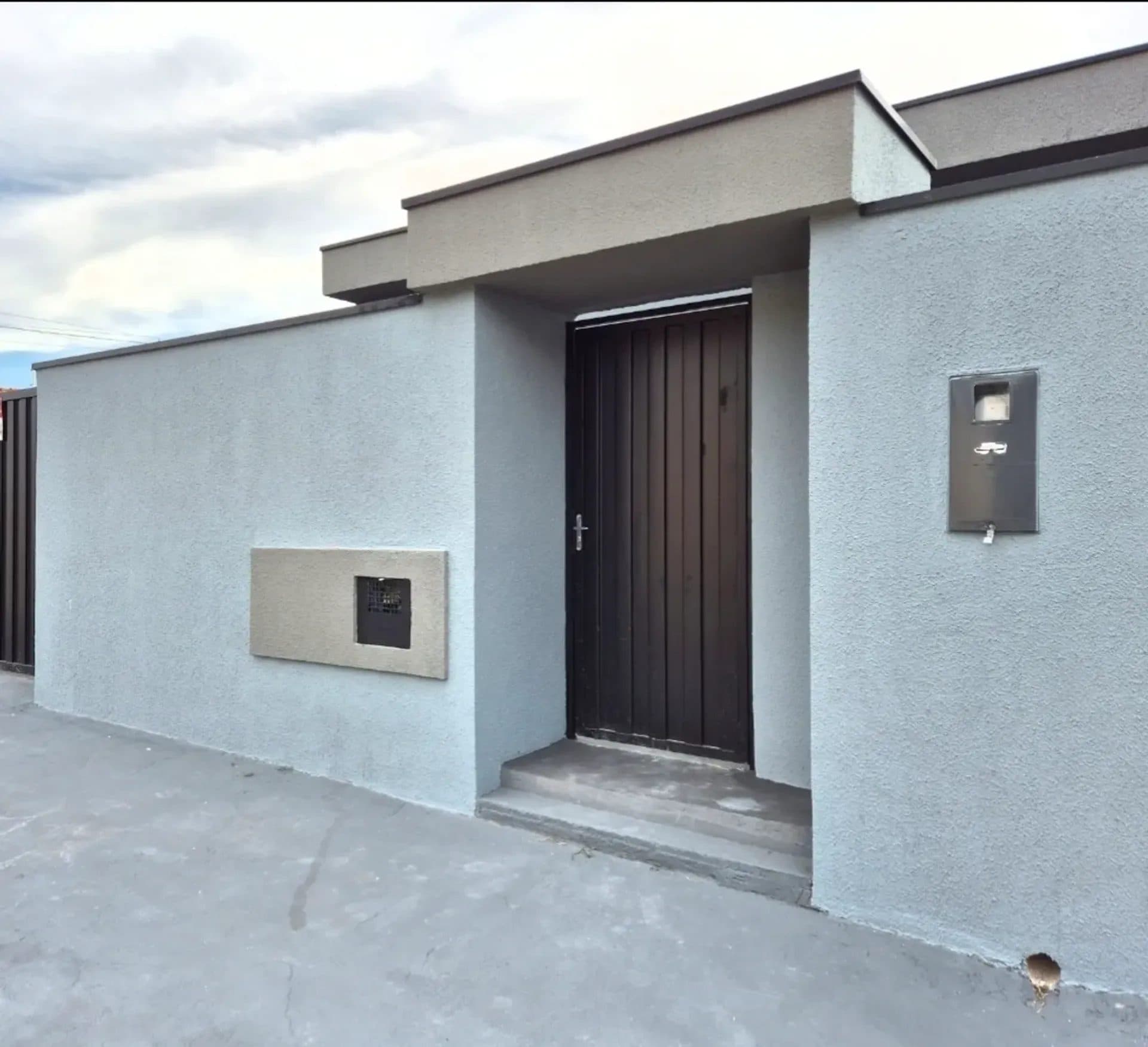Fachada frontal da casa com muro cinza, portão escuro e piso de concreto.