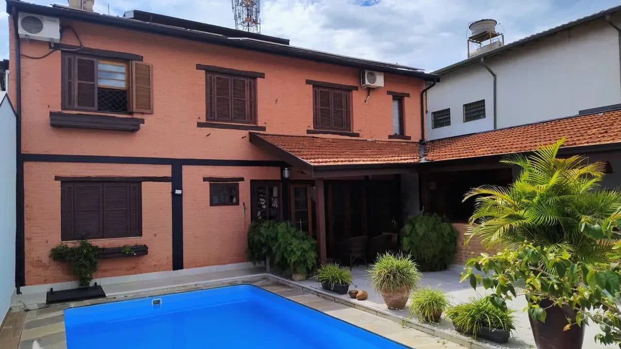 Fachada dos fundos com piscina azul, parede em tom avermelhado e janelas com venezianas de madeira.