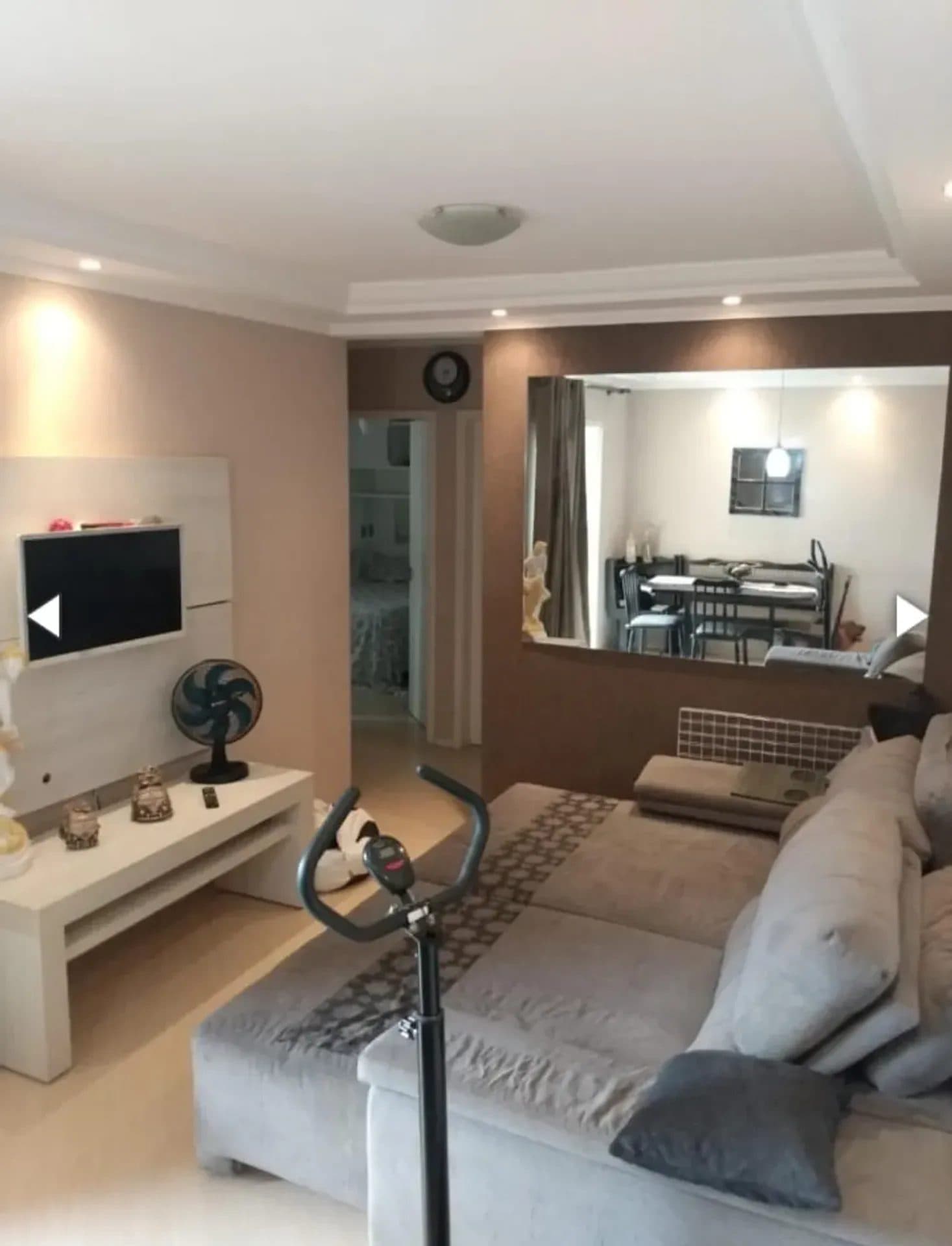 Sala de estar com sofá cinza, rack branco, TV e espelho na parede.